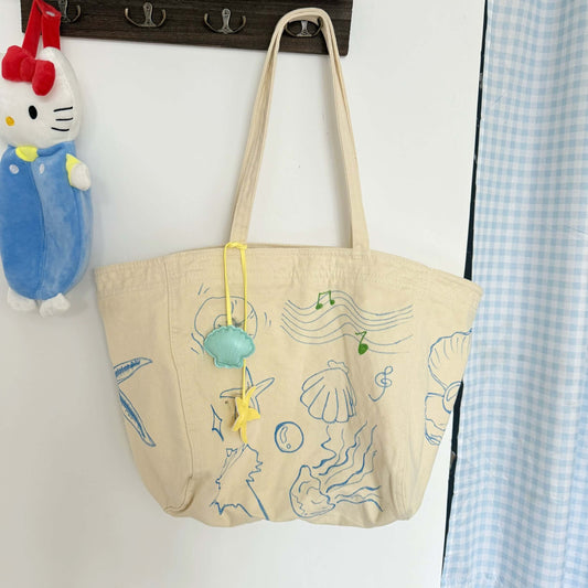 Beige tote bag with mermaid design hanging on a hook next to a Hello Kitty plush toy.