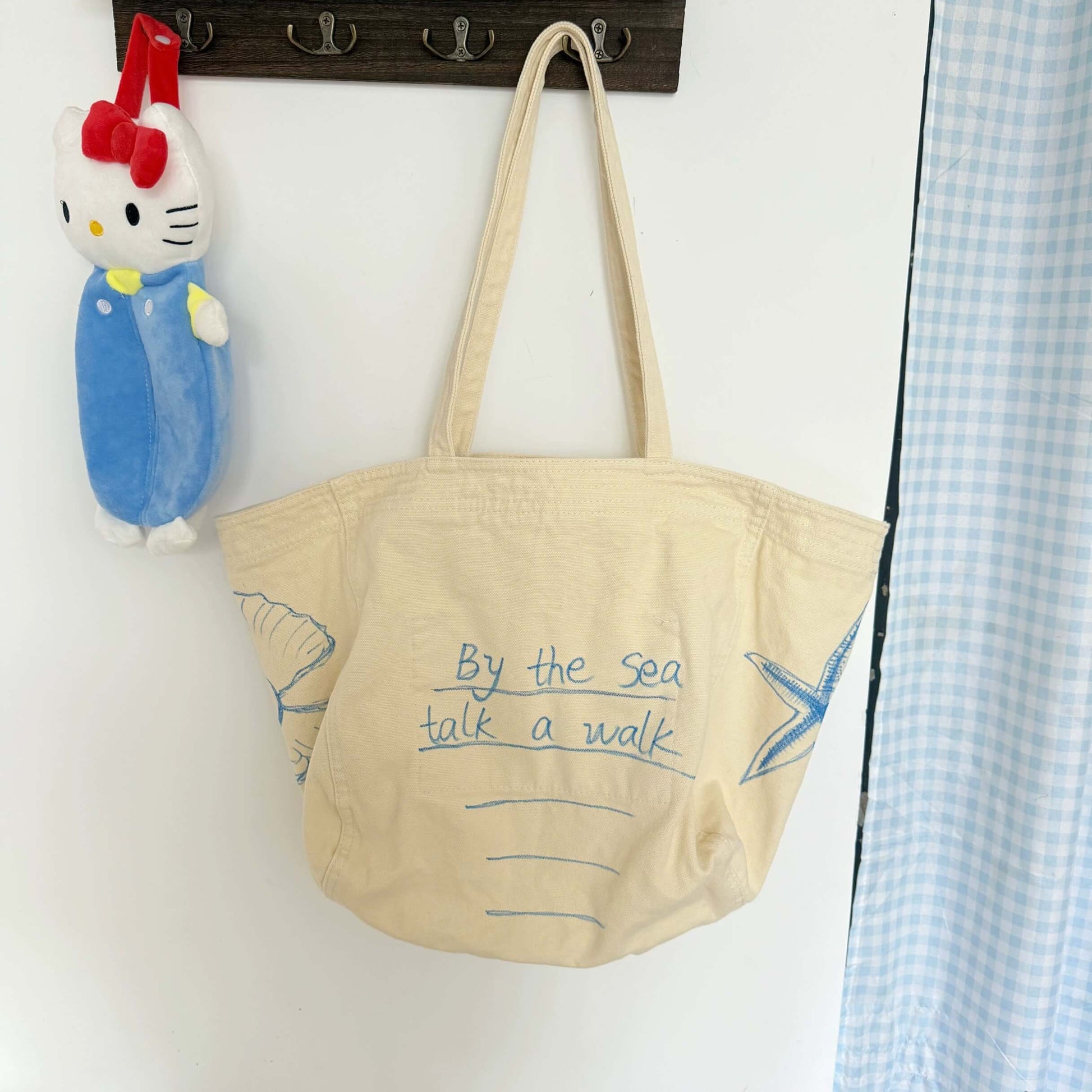 Beige tote bag with text and starfish design hanging on a wall next to a Hello Kitty toy.