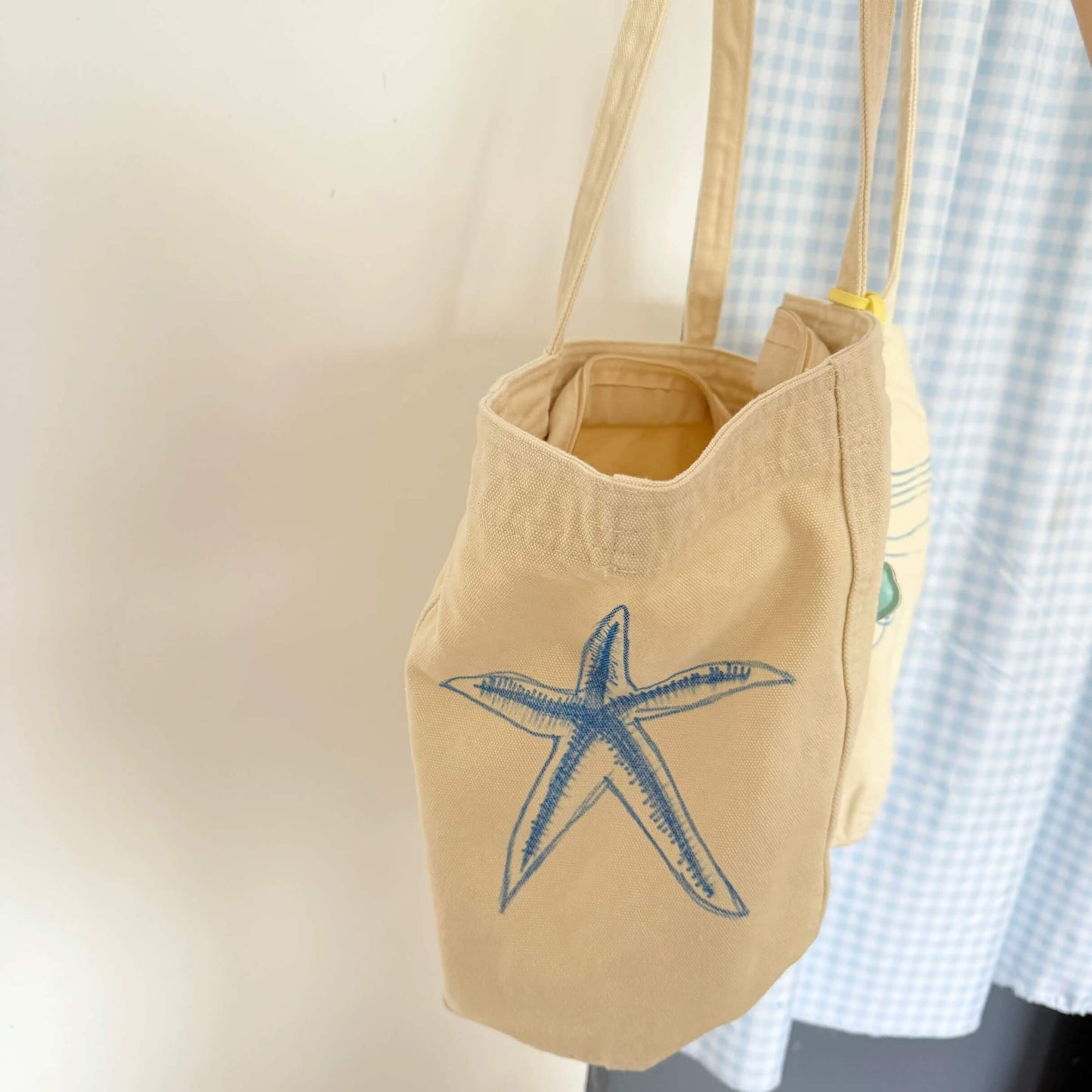 Beige tote bag with a blue starfish design hanging on a mannequin against a neutral background.